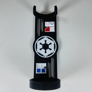 May include: Black stand with a circular base and a curved top. The stand features a white and black emblem in the center, with smaller rectangular designs above and below. The design includes red and blue accents.