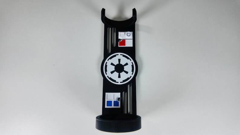May include: Black stand with a circular base and a curved top. The stand features a white and black emblem in the center, with smaller rectangular designs above and below. The design includes red and blue accents.