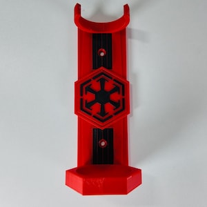 Puede incluir: Un soporte de pared rojo impreso en 3D para sable de luz. El soporte presenta un símbolo imperial negro y rojo en el centro. El soporte tiene una parte superior curva y una base hexagonal. El soporte está diseñado para sostener un sable de luz.