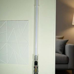 May include: A lightsaber with a silver handle and a white blade is mounted on a wall. The handle has black and red accents with a clear emitter. The lightsaber is a prop from the Star Wars franchise.