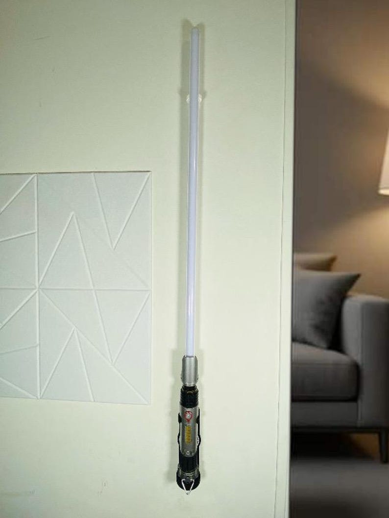 May include: A lightsaber with a silver handle and a white blade is mounted on a wall. The handle has black and red accents with a clear emitter. The lightsaber is a prop from the Star Wars franchise.