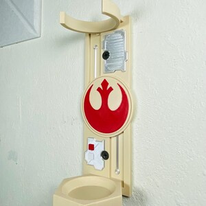 May include: A beige wall-mounted holder featuring a red and white emblem. The holder has a curved top, a small shelf, and several decorative elements. The design includes a circular emblem with a red symbol.