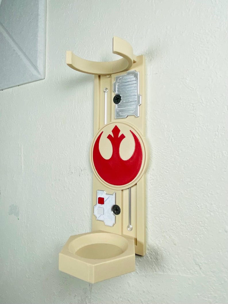 May include: A beige wall-mounted holder featuring a red and white emblem. The holder has a curved top, a small shelf, and several decorative elements. The design includes a circular emblem with a red symbol.