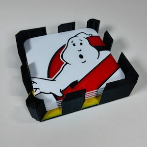 May include: A set of square coasters featuring the Ghostbusters logo, with a white ghost figure and red stripe on a white background. The coasters are held in a black and yellow holder.