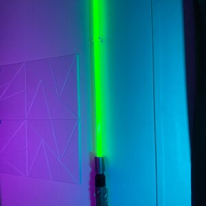 May include: A green lightsaber with a silver and black handle is mounted on a wall. The lightsaber's blade glows brightly, and the handle has intricate details. The wall is painted in shades of blue and purple.