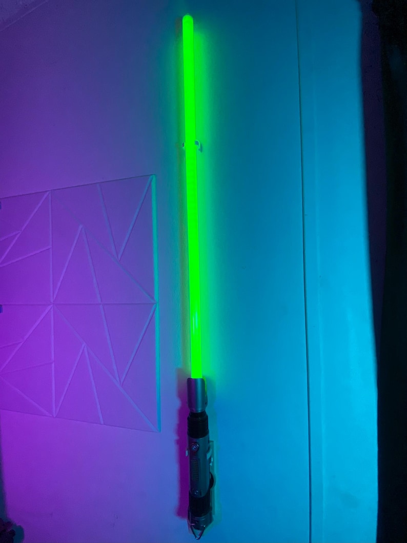May include: A green lightsaber with a silver and black handle is mounted on a wall. The lightsaber's blade glows brightly, and the handle has intricate details. The wall is painted in shades of blue and purple.