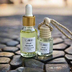 May include: Two glass bottles of Calido Citron Mandarin car diffuser refill. One bottle has a gold cap with a white dropper, the other has a wooden cap and a string. The bottles are filled with a yellow liquid and sit on a stone surface.