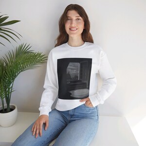 May include: White long-sleeve sweatshirt featuring a black and white image of a window with a view of a city. The text in the image is partially visible. The sweatshirt is paired with blue jeans.