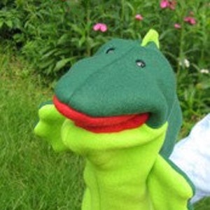 May include: A green and lime green felt hand puppet shaped like a friendly alligator. The alligator has a red mouth, a green head, and lime green arms and legs. The puppet is set against a background of green grass and foliage.