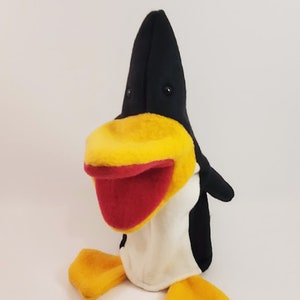 May include: Black and white penguin puppet with a yellow beak and red mouth. The puppet has a white belly and yellow feet.