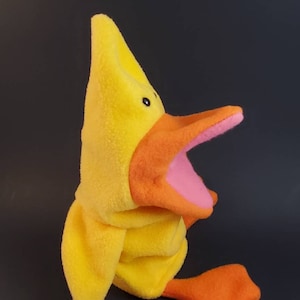 May include: Yellow plush hand puppet shaped like a duck with an orange beak and pink inside the mouth. The duck has black eyes and is sitting on a black surface.