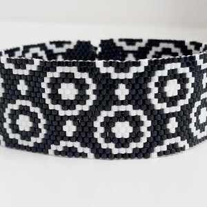 May include: Black and white beaded bracelet with a geometric pattern. The bracelet is made of small, square beads that are arranged in a repeating design of circles and squares.
