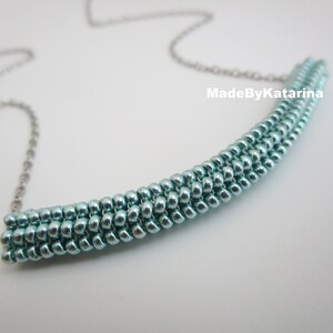 Può includere: Una collana con catena d'argento con un ciondolo a barra di perline blu chiaro. Il ciondolo è fatto di piccole perline rotonde disposte in linea retta. Il testo "MadeByKatarina" è visibile nell'immagine.