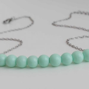 May include: A delicate necklace featuring a silver chain and a row of ten light-green, round beads. The beads are evenly spaced along the chain, creating a minimalist and elegant design. The necklace is displayed against a white background.