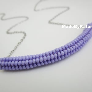 Può includere: Una collana di perline lavanda con un design curvo. La collana presenta una serie di piccole perline rotonde in una tonalità viola chiaro, infilate insieme per formare una barra curva. La collana è attaccata a una catena d'argento. Il testo "MadeByKatarina" è visibile.