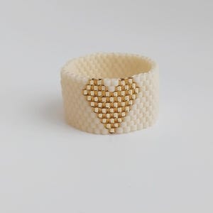 May include: A wide, ivory-colored beaded ring with a gold heart design. The heart is centered on the ring and is made of small, gold beads. The ring is approximately 1 inch wide.