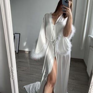 May include: White satin robe with long sleeves and a shawl collar. The robe has a front closure and a belt. The sleeves and collar are trimmed with white feather-like material. The robe is floor-length and has a side slit.  The luxurious robe is perfect for relaxing at home.