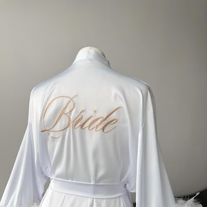 May include: White satin robe with feather trim on the sleeves. The robe has a sash and the words 'Bride' embroidered on the back in gold thread.