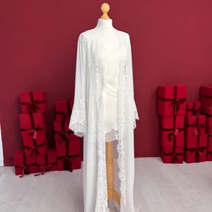 Pode incluir: Um roup&atilde;o e um vestido branco, expostos em um manequim. O roup&atilde;o apresenta delicadas bordas de renda nas bordas e mangas. O fundo &eacute; vermelho com v&aacute;rias caixas de presente vermelhas.