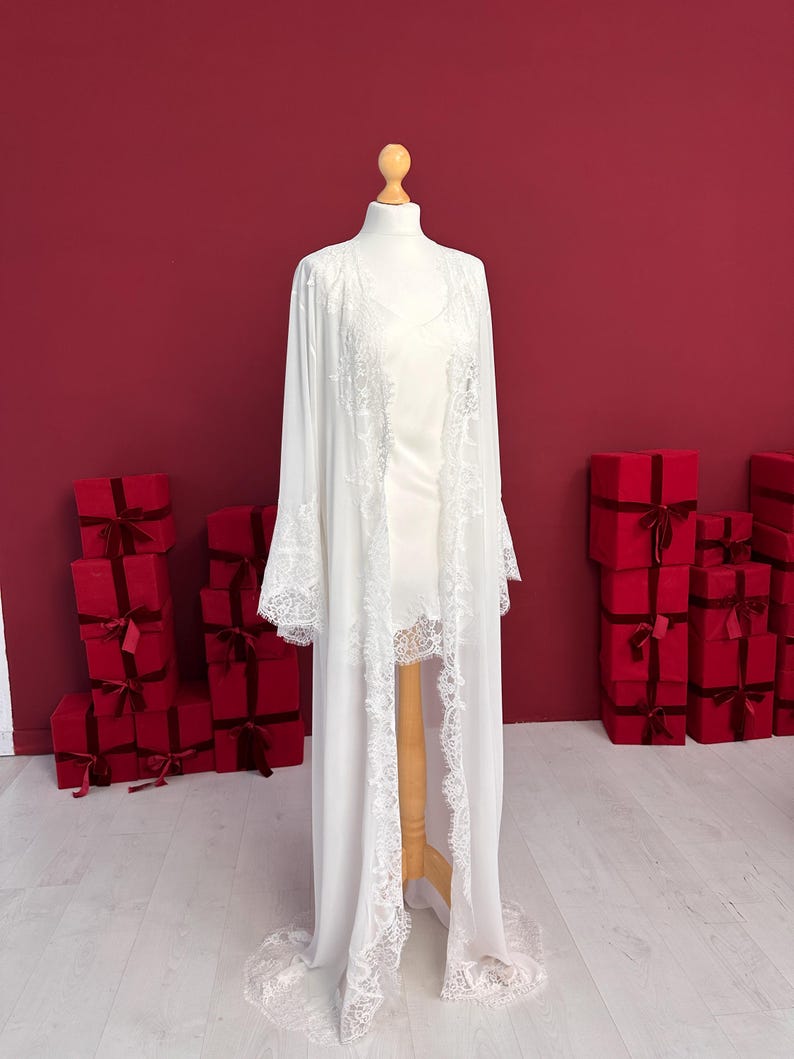 Pode incluir: Um roup&atilde;o e um vestido branco, expostos em um manequim. O roup&atilde;o apresenta delicadas bordas de renda nas bordas e mangas. O fundo &eacute; vermelho com v&aacute;rias caixas de presente vermelhas.