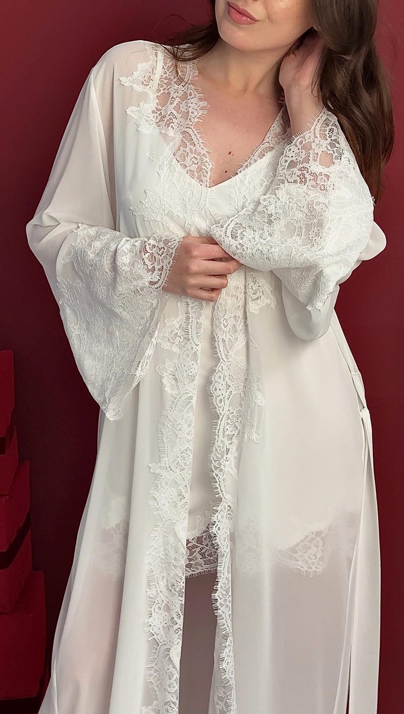 Pode incluir: Conjunto de lingerie branco com um roup&atilde;o transparente com acabamento em renda. O roup&atilde;o tem mangas compridas com punhos largos de renda e um acabamento em renda a condizer na frente e na bainha. O conjunto inclui uma camisola e cal&ccedil;&otilde;es, tamb&eacute;m com detalhes em renda.
