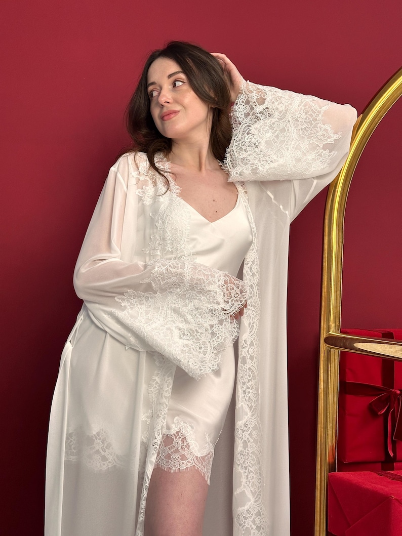 Pode incluir: Um conjunto de lingerie de cetim branco, incluindo um vestido de al&ccedil;a e um roup&atilde;o longo com acabamento em renda. O roup&atilde;o tem mangas largas com punhos de renda. A mulher est&aacute; encostada a uma moldura dourada, com fundo vermelho.
