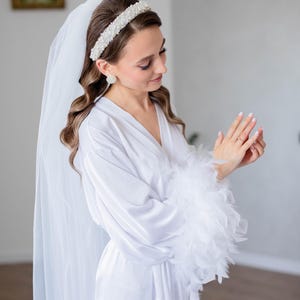 May include: A white satin robe with feather trim on the sleeves. The robe is worn by a person with a white veil and a pearl headband.
