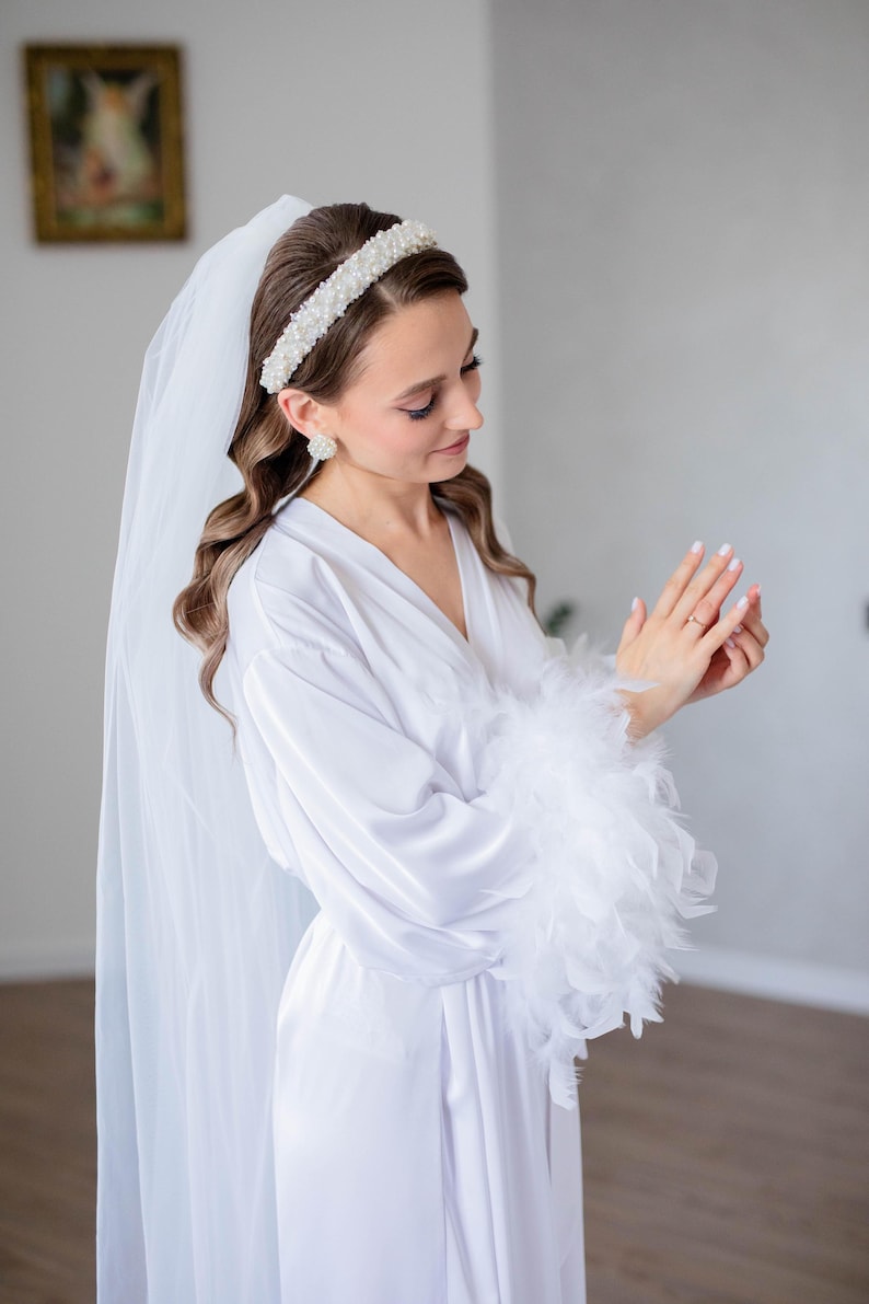 May include: A white satin robe with feather trim on the sleeves. The robe is worn by a person with a white veil and a pearl headband.