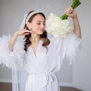May include: A white satin robe with feather trim on the sleeves. The robe is tied at the waist with a matching sash. The person is wearing a white headband with pearls and pearl earrings.