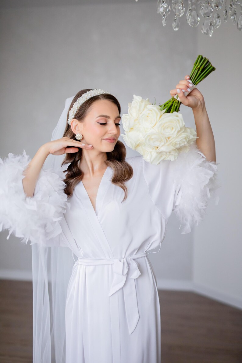 May include: A white satin robe with feather trim on the sleeves. The robe is tied at the waist with a matching sash. The person is wearing a white headband with pearls and pearl earrings.