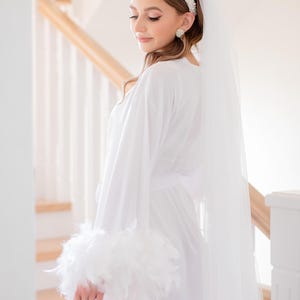 May include: A white satin robe with feather trim on the sleeves. The robe is worn by a person with a white veil and a pearl headband. The person is standing in front of a white staircase.