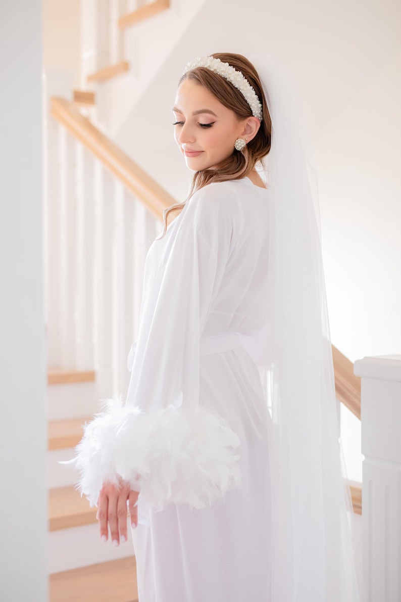 May include: A white satin robe with feather trim on the sleeves. The robe is worn by a person with a white veil and a pearl headband. The person is standing in front of a white staircase.