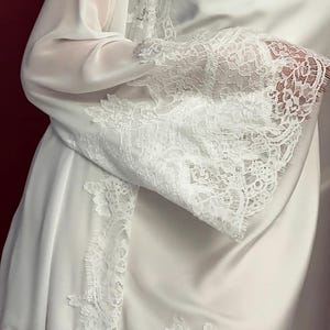 Pode incluir: Robe e vestido de cetim branco com delicadas rendas. O robe apresenta mangas transparentes e detalhes de renda nas bordas. O vestido tem decote em V. O visual geral &eacute; elegante e feminino.