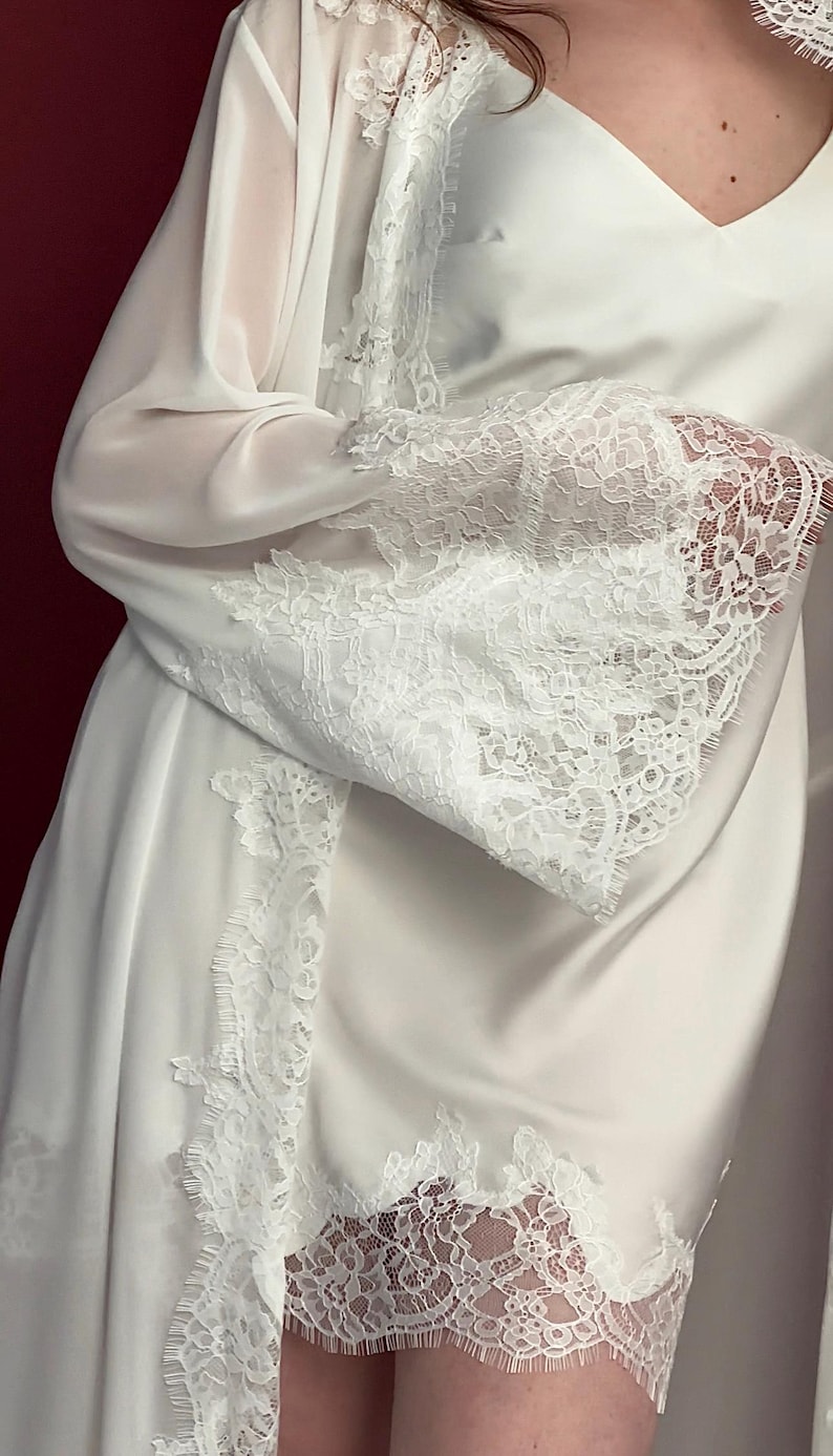 Pode incluir: Robe e vestido de cetim branco com delicadas rendas. O robe apresenta mangas transparentes e detalhes de renda nas bordas. O vestido tem decote em V. O visual geral &eacute; elegante e feminino.