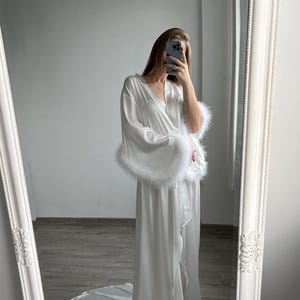 May include: White satin robe with long sleeves and a floor-length silhouette. The sleeves have white feather trim. The robe has a wrap style and is shown in a full-length view.