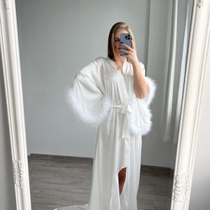 May include: White satin robe with long sleeves and a floor-length hem. The sleeves have white feather trim.  The robe has a self-tie belt and a front slit.  It's a luxurious, elegant style perfect for bridal or special occasions.