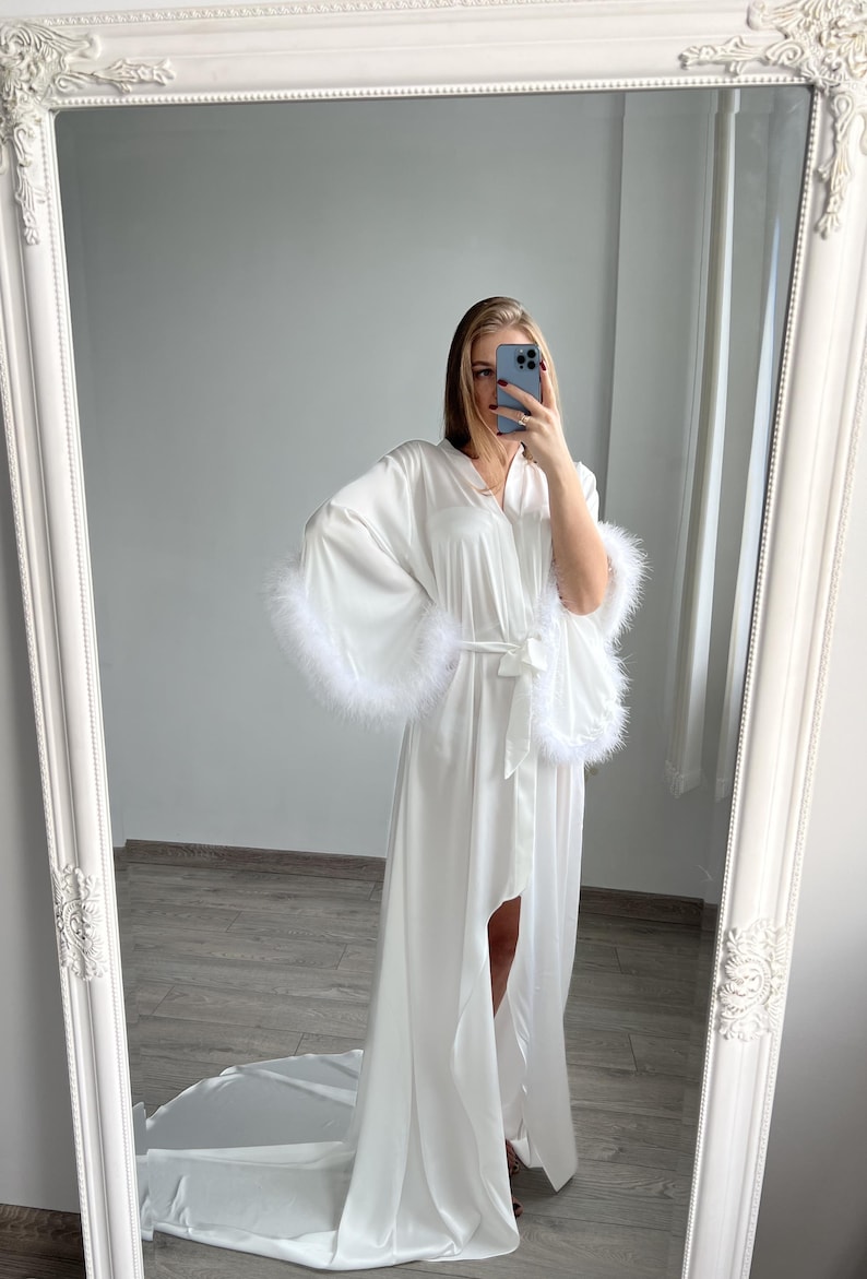 May include: White satin robe with long sleeves and a floor-length hem. The sleeves have white feather trim.  The robe has a self-tie belt and a front slit.  It's a luxurious, elegant style perfect for bridal or special occasions.