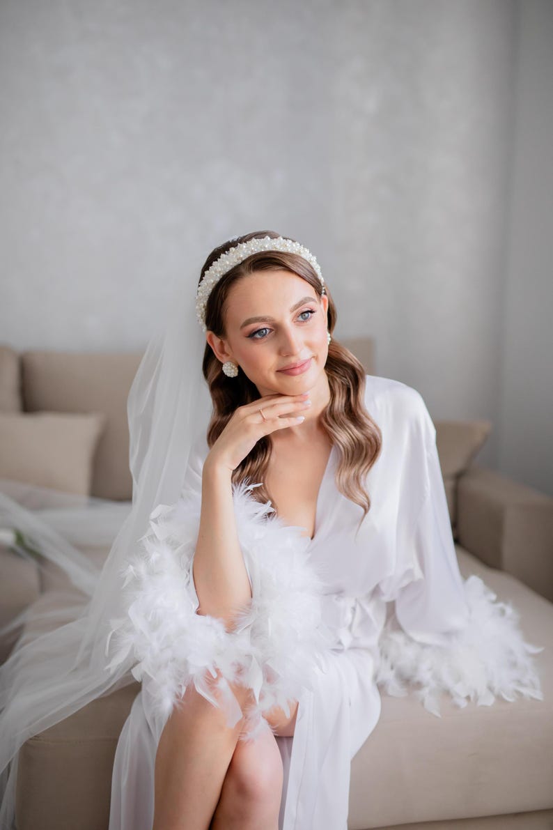 May include: A bride wearing a white satin robe with feather trim and a pearl headband.