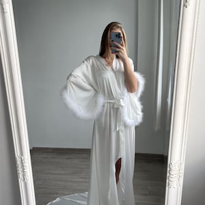 May include: White satin robe with long sleeves and feather trim. The robe has a long, flowing design and a belted waist. It is shown in a full-length view, displayed against a light-colored background.