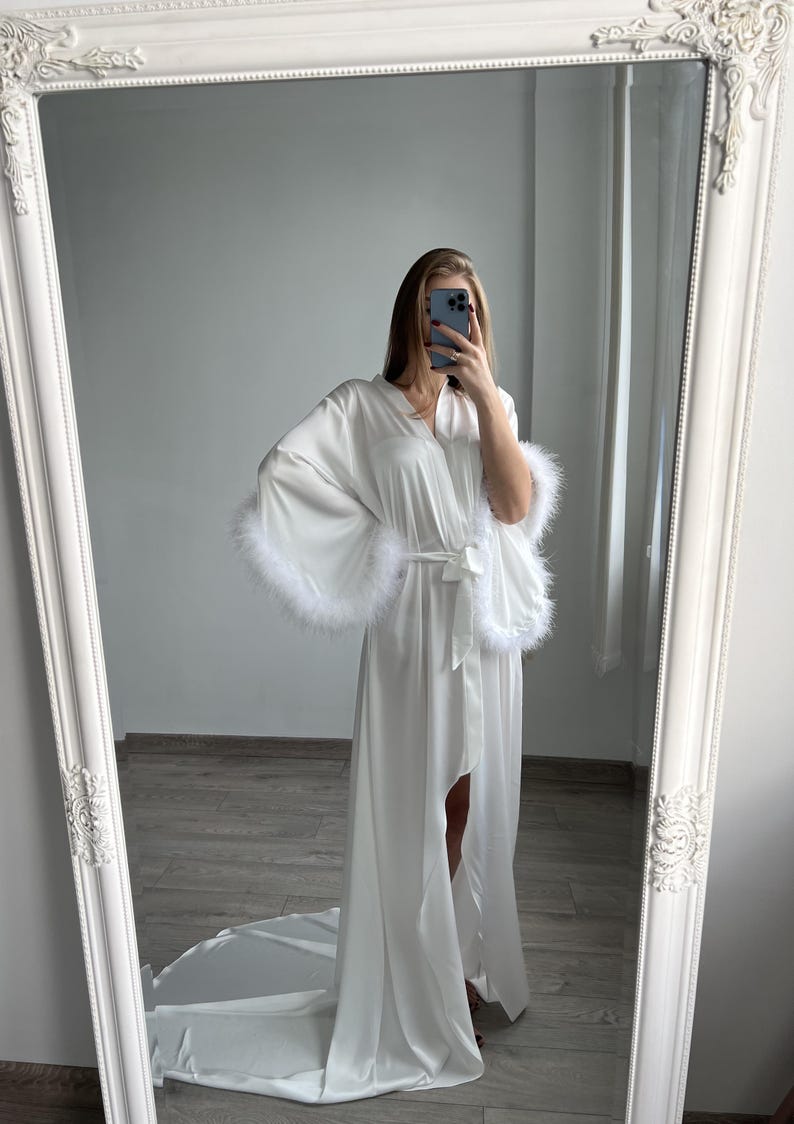 May include: White satin robe with long sleeves and feather trim. The robe has a long, flowing design and a belted waist. It is shown in a full-length view, displayed against a light-colored background.