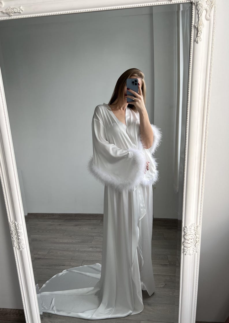 May include: White satin robe with long sleeves and a floor-length silhouette. The sleeves have white feather trim. The robe has a wrap closure and a relaxed fit. The luxurious fabric drapes elegantly. This elegant robe is perfect for bridal boudoir photos or a relaxing evening at home.