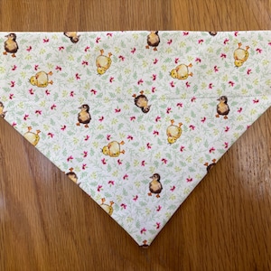 Puede incluir: Bandana blanca para mascotas con un estampado repetido de patitos marrones y amarillos, pequeñas flores rojas y hojas verdes. La bandana está doblada en forma de triángulo.