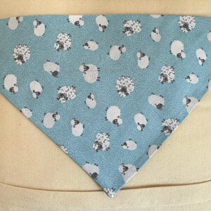 Puede incluir: Bandana triangular azul claro para mascotas con un patrón repetido de ovejas blancas y grises. Las ovejas están representadas en varias poses y tamaños. La bandana está hecha de tela suave.