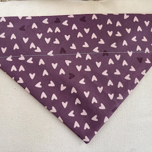 Puede incluir: Bandana triangular para mascotas en tela morada oscura, estampada con pequeños corazones rosa claro. La bandana está doblada y tiene un dobladillo cosido en blanco en el borde superior. La tela es suave y probablemente de algodón.