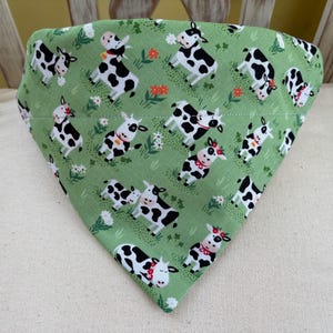 Puede incluir: Un pañuelo triangular verde para mascotas con un patrón repetido de vacas de dibujos animados. Las vacas tienen manchas negras, lazos rojos y detalles florales. El fondo presenta hierba verde y flores blancas y naranjas.
