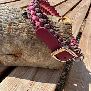 Puede incluir: Un collar trenzado para perros en tonos rosa y gris, con una correa de cuero burdeos y una hebilla y un anillo de metal plateado. El collar se muestra sobre un trozo de madera.