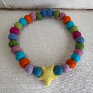 Puede incluir: Un collar colorido con un colgante de estrella amarilla y una cadena de bolas de fieltro multicolores. El collar presenta bolas azules, naranjas, verdes y rosas. El artículo se muestra sobre un fondo neutro.
