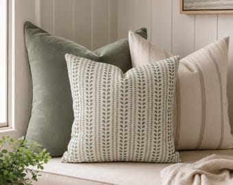 Può includere: Tre cuscini quadrati disposti su una panca. Uno è verde salvia a tinta unita, uno è color crema con motivi a foglie verticali verde salvia e uno è color crema con sottili righe verticali. Una coperta color crema con frange è appoggiata vicino.