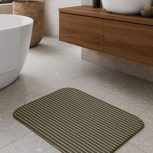Op de afbeelding: Een rechthoekige olijfgroene badmat met lichtbeige strepen. De mat ligt op een lichtgrijze tegelvloer in een moderne badkamer. De afgeronde hoeken van de mat verzachten het ontwerp. Afmetingen ca. 70 cm x 50 cm.