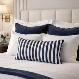 Op de afbeelding: Een bed met wit beddengoed en verschillende kussens. Er zijn twee marineblauwe fluwelen kussens, twee witte kussens en een gestreept kussen met marineblauwe en witte strepen. Aan het voeteneinde van het bed ligt een marineblauwe loper.